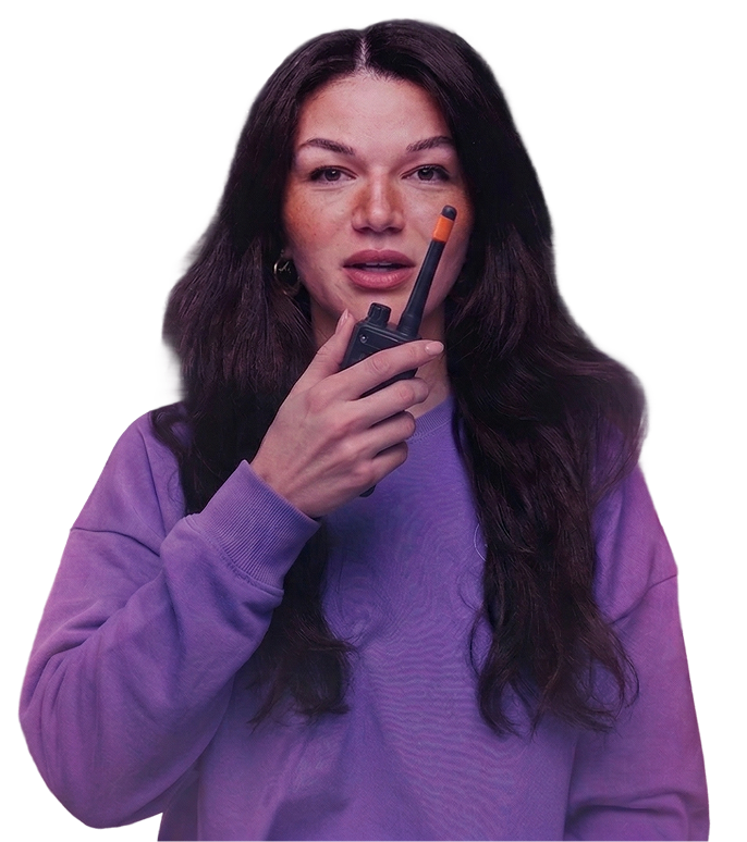 Eine Frau mit dunklen Haaren trägt einen lila Pullover und hält ein Funkgerät an ihren Mund.